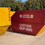 Red skip with white 'LOSTOCK SKIP HIRE' lettering, set between yellow containment barriers outdoors.