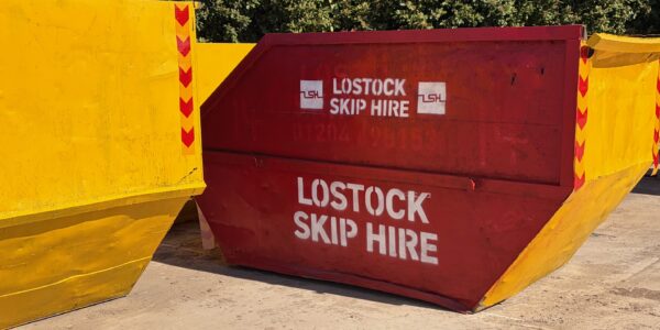 Red skip with white 'LOSTOCK SKIP HIRE' lettering, set between yellow containment barriers outdoors.