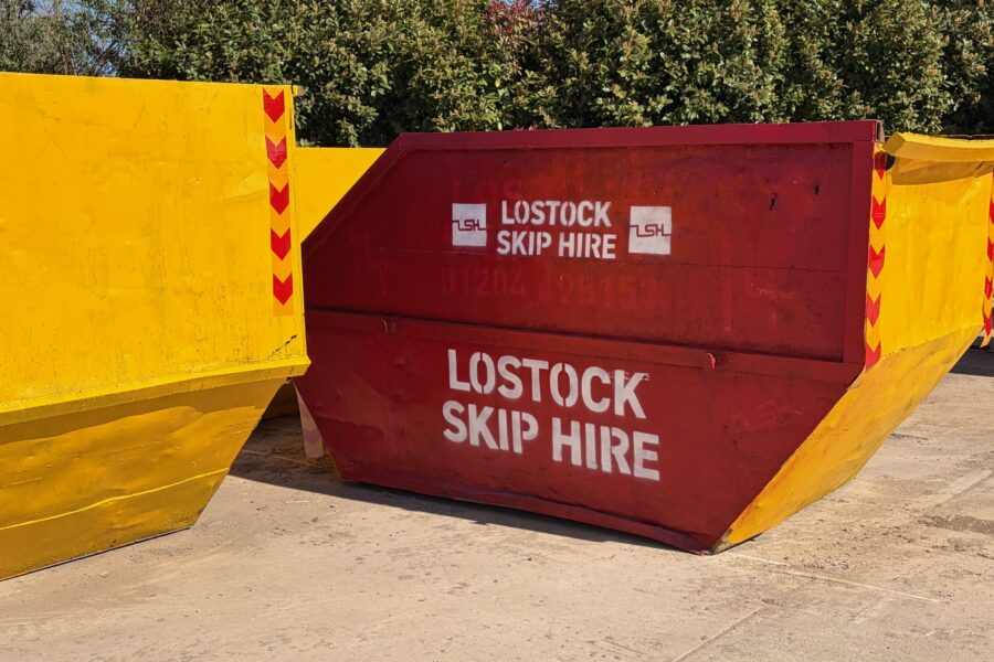 Red skip with white 'LOSTOCK SKIP HIRE' lettering, set between yellow containment barriers outdoors.