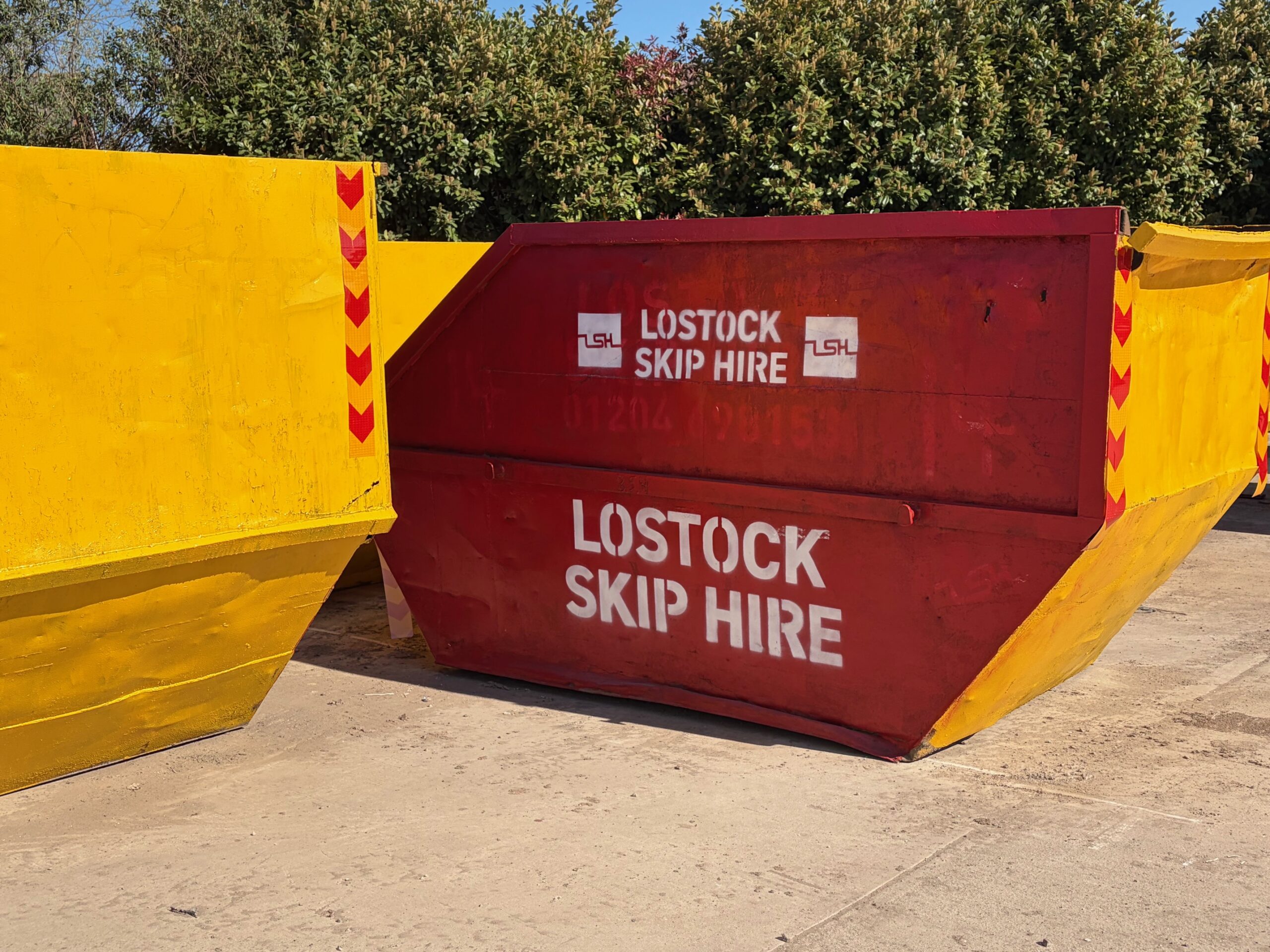 Red skip with white 'LOSTOCK SKIP HIRE' lettering, set between yellow containment barriers outdoors.