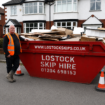 Worker in orange hi-vis vest beside a red skip filled with cardboard on a residential street