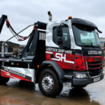 Tow truck with hydraulic arms and 'Lostock Skip Hire' branding in a scrapyard with piles of rubble behind it
