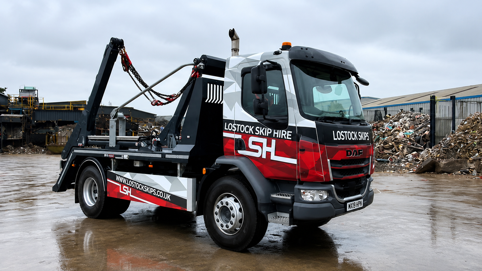 Tow truck with hydraulic arms and 'Lostock Skip Hire' branding in a scrapyard with piles of rubble behind it