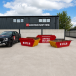 Black pickup truck with Lostock Skip Hire logo parked beside red and yellow skips in a paved lot with the company building in the background.