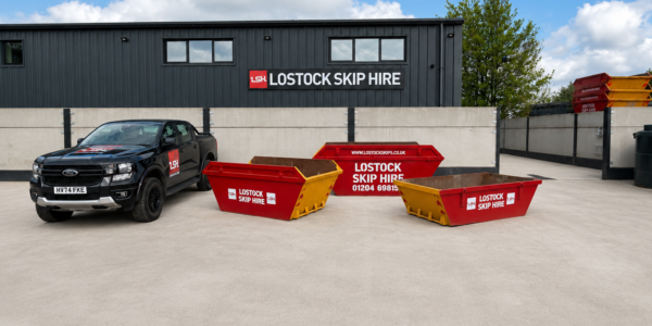Black pickup truck with Lostock Skip Hire logo parked beside red and yellow skips in a paved lot with the company building in the background.