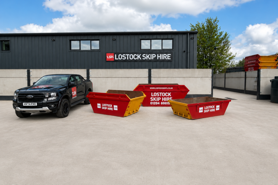 Black pickup truck with Lostock Skip Hire logo parked beside red and yellow skips in a paved lot with the company building in the background.