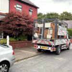 Skip hire truck with 'Lostock Skip Hire' graphics parked on a residential street near a red-leaved tree and shopfronts.