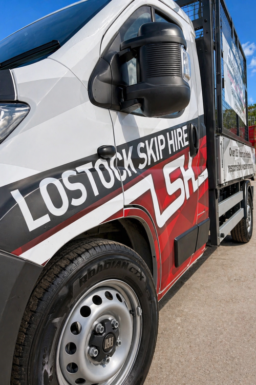 White cargo van with bold 'LOSTOCK SKIP HIRE' branding along the side, close-up of front wheel and side mirror, bright blue sky in background.
